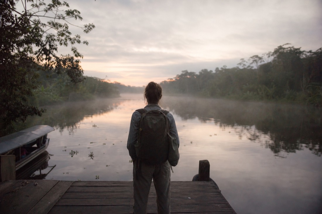 Traveler with a backpack looking out over a jungle river from a wooden dock at first light