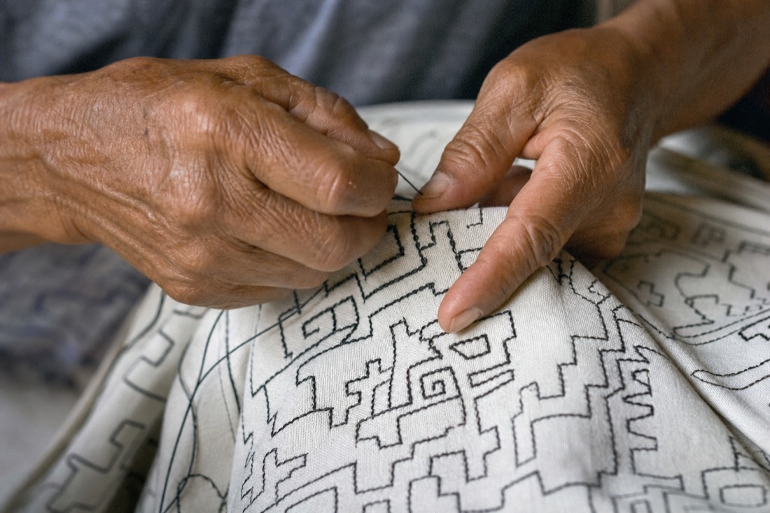 Elderly hands weaving a traditional Shipibo geometric pattern on white cloth
