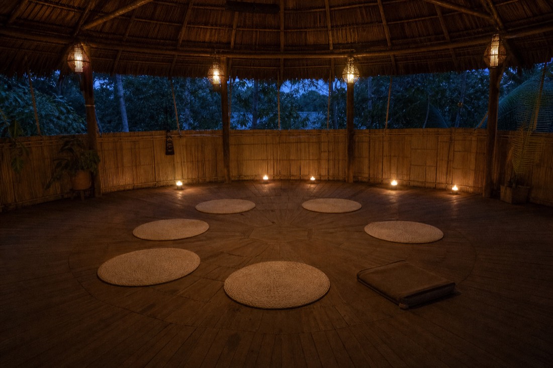 Small circle of mats inside a softly lit maloca, a facilitator's seat close to the guests