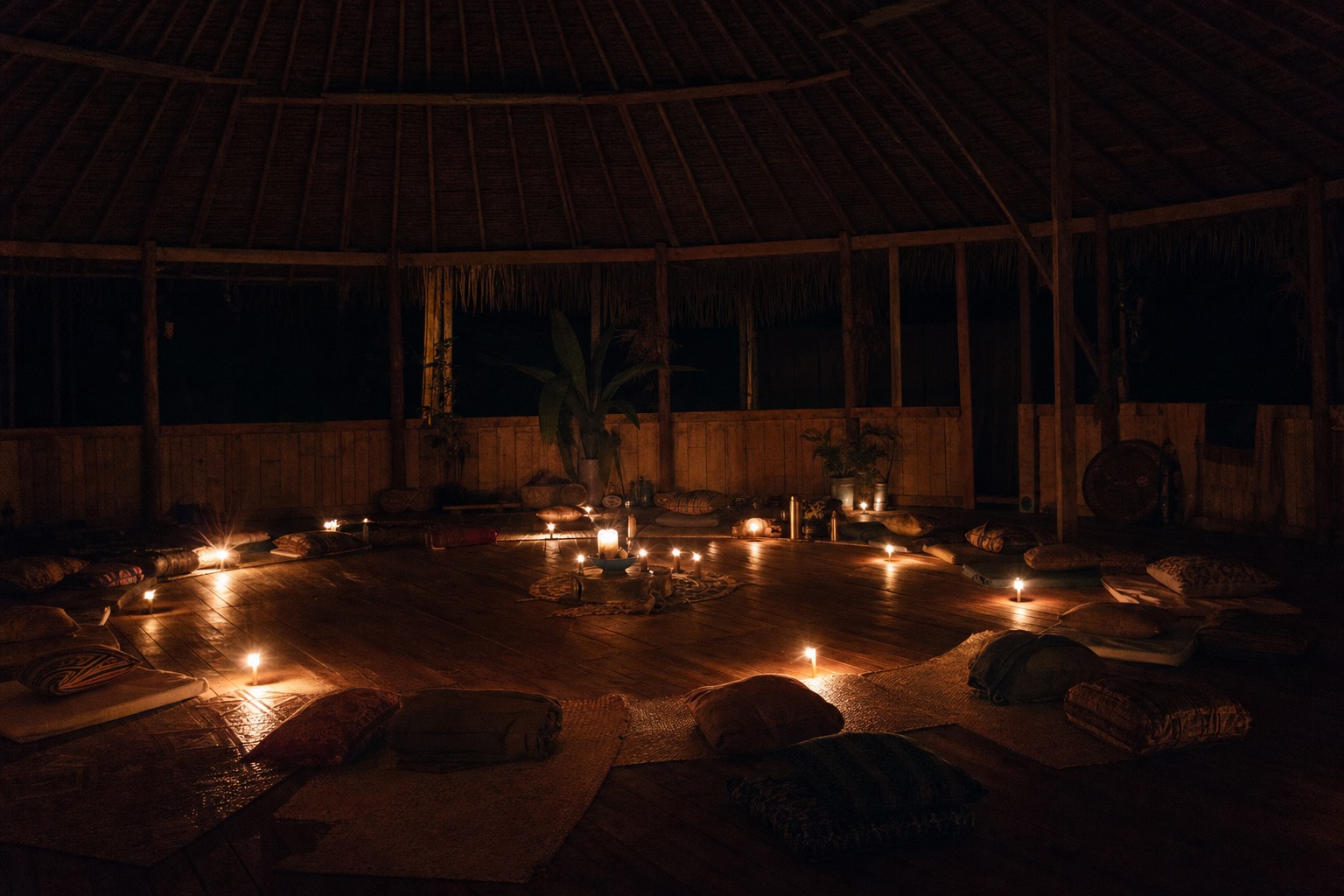 Empty ceremony maloca at night lit by candles, cushions arranged in a circle