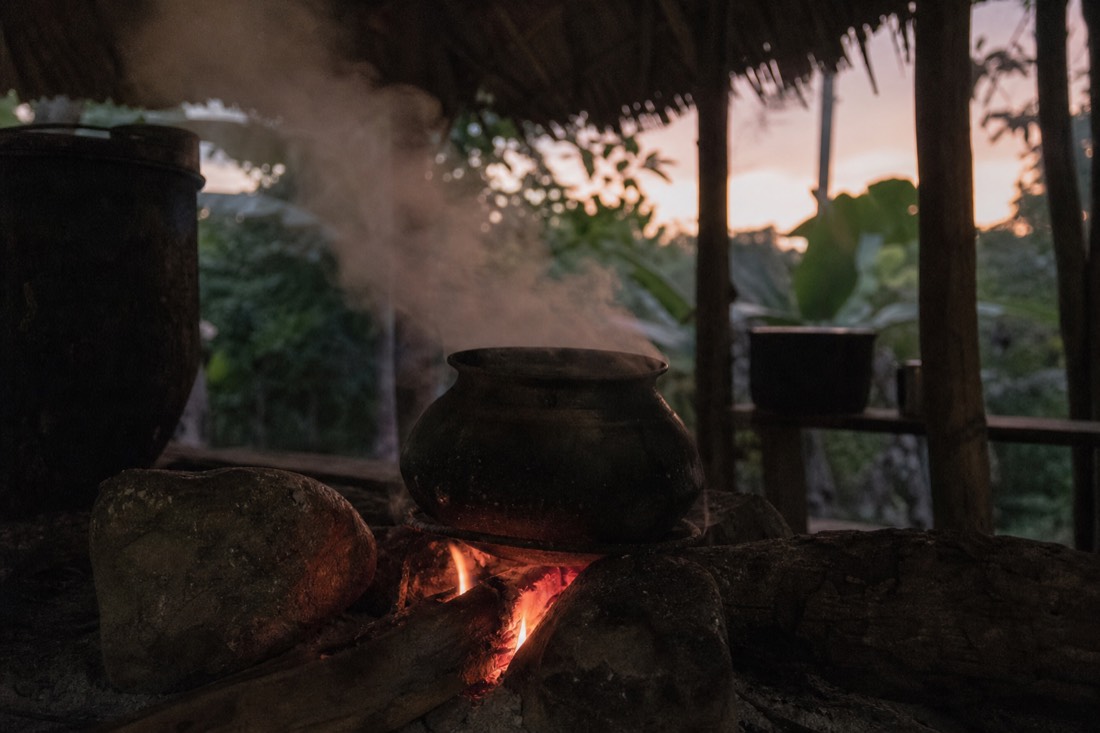 Close-up of steam rising from a clay pot over a fire in a jungle cooking hut at dusk