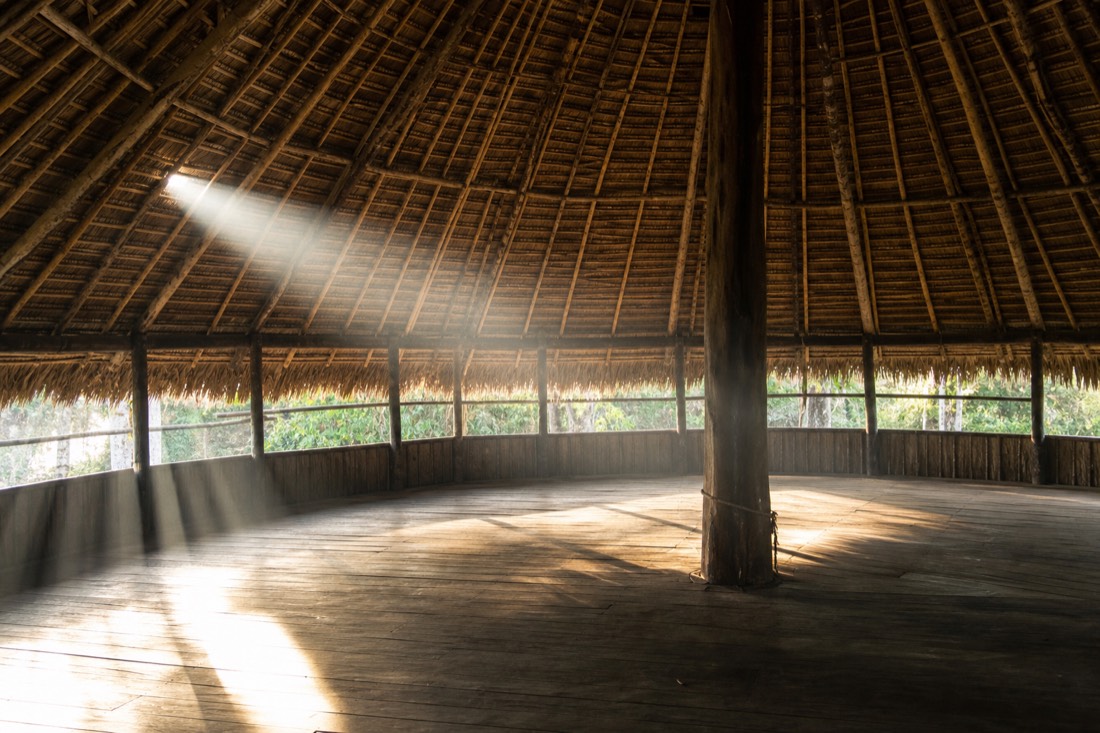 Empty maloca interior in daylight with sunbeams crossing the wooden floor toward the center pole