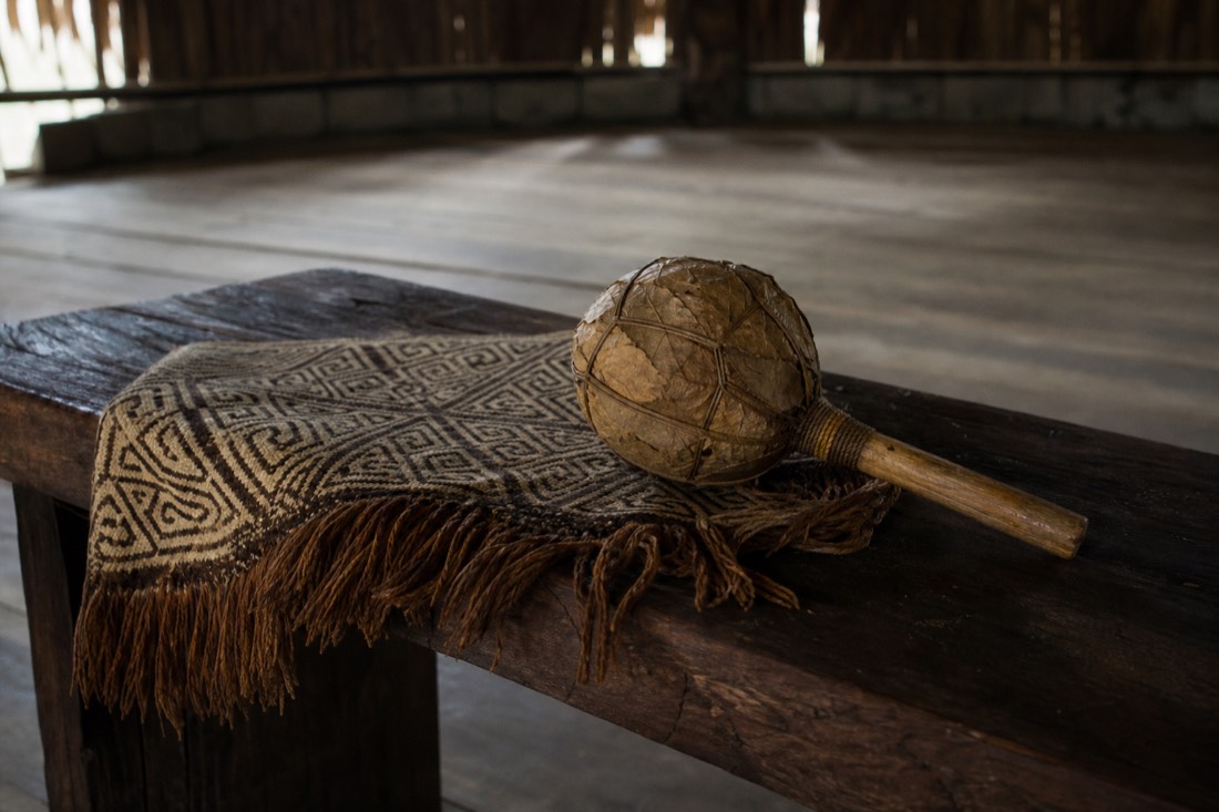Woven Shipibo-patterned textile and a leaf rattle resting on a wooden bench inside an empty ceremonial space